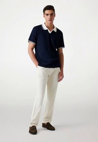 Marineblaues Poloshirt mit weißem Kragen und weißen Manschetten, kombiniert mit cremefarbenen Hosen und braunen Loafers. Glatter Stoff, entspannte Passform, schlichtes Design.
