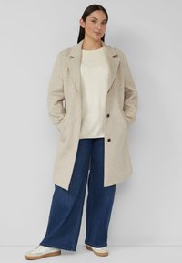 Vrouw draagt een beige jas, crème trui, wijde blauwe spijkerbroek en witte sneakers, staand met handen in de jaszakken tegen een effen achtergrond.