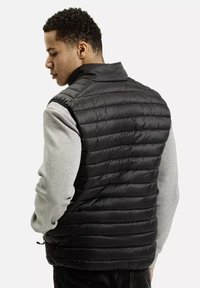 Czarna kamizelka puchowa z poziomym pikowaniem, wysokim kołnierzem i zapięciem na zamek, zestawiona z szara koszulką z długim rękawem pod spodem.