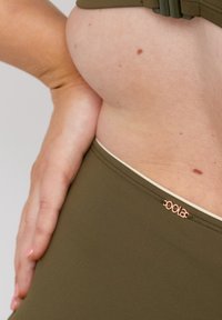 Olivgrön badkläder med en mjuk textur, som har en roséguld logo-accent nära midjebandet. Närbild av midjan och handposition.