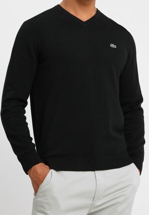 Mand iført en sort V-hals sweater med en lille krokodille-logo på brystet og lyse bukser, hænderne i lommerne.