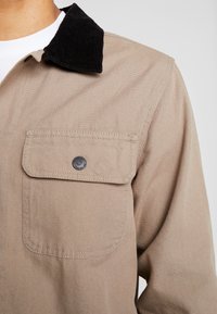 Beige takki mustalla samettikauluksella, vasemmassa rinnassa läpällä varustettu Tasku mustalla nepparilla, päällä valkoinen pyöreäkaula-aukkoinen paita.