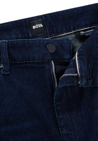 Mörkblå slim fit denimjeans med delvis öppen dragkedja, svart knapp och etiketter som läser "BOSS" och "DELAWARE."