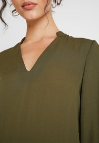 Maglietta verde oliva con scollo a V e texture morbida e liscia. Presenta una piega sottile al collo e maniche lunghe, leggermente arricciate.