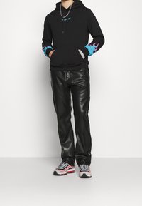 Haut à capuche noir avec des accents de flammes bleues et roses sur les poignets, associé à un pantalon en cuir noir et des baskets gris-rouge avec des détails en mesh.