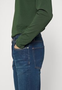 Tmavě zelený dlouhý rukáv s přiléhavými rukávy v kombinaci s modrými džínami, které mají švovou kapsu a jemné vyblednutí na látce.