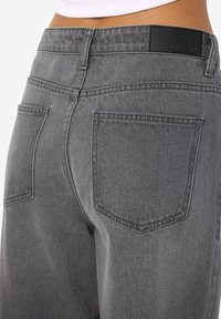 Lähikuva henkilöstä, joka pukeutunut korkeavyötäröisiin harmaisiin farkkuihin, joissa on kaksi takataskua ja musta vyötäröetiketti.