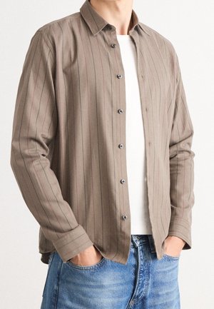 Mann trägt ein aufgeknöpftes beiges Nadelstreifenhemd über einem weißen T-Shirt und hat die Hände in den Vordertaschen einer ausgewaschenen blauen Jeans.