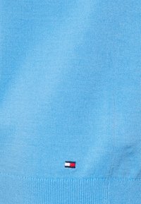 Světle modrý pletený svetr s žebrovaným spodním lemem a malou nášivkou loga s červenými, bílými a modrými pruhy blízko levého dolního rohu.