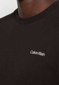 Gros plan sur une personne portant un t-shirt noir à col rond avec un petit logo blanc Calvin Klein sur la poitrine.