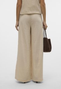 Vrouw staand, gekleed in beige broek met wijde pijpen en top, met een kleine bruine suède handtas in haar rechterhand.