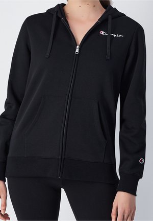 Czarna bluza z kapturem zapinana na zamek, z przednimi kieszeniami, kapturem ze sznurkiem oraz logo Champion na piersi i lewym rękawie, noszona na czarnych legginsach.