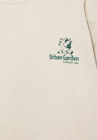 Tissu beige avec logo imprimé en vert représentant un bol de fruits au-dessus du texte « Urban Garden » et un texte plus petit « Lifestyle stay » sur la zone de la poitrine.