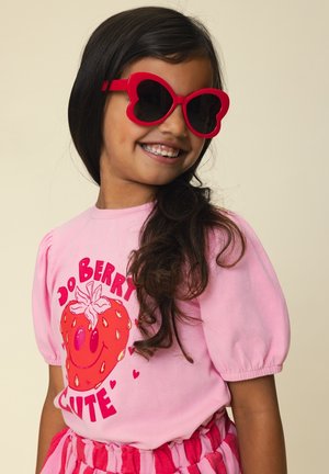 Lachend meisje met lang donker haar, droeg rode hartvormige zonnebril en een roze shirt met een aardbei en de tekst "Do Berry Cute".