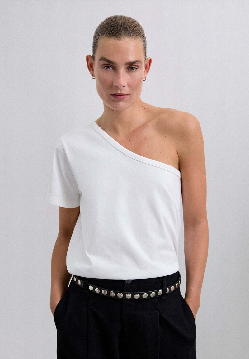 Weißes T-Shirt mit kurzen Ärmeln, asymmetrischem Ausschnitt und lockerer Passform. Kombiniert mit hoch taillierten schwarzen Hosen und einem Nieten-Gürtel.