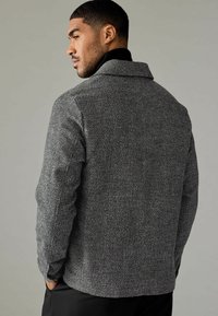 Jachetă texturată gri cu guler, design ajustat și modele subtile, având o croială strânsă și mâneci lungi, prezentată dintr-un unghi lateral.