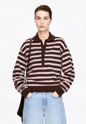 Woman with tied-back hair wearing a brown, pink, and blue striped sweater with buttons, light blue jeans, and a black shoulder bag.