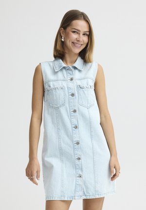 Lachende vrouw met schouderlang haar, gekleed in een lichtblauwe, mouwloze spijkerjurk met knopen en zakken, tegen een egale achtergrond.