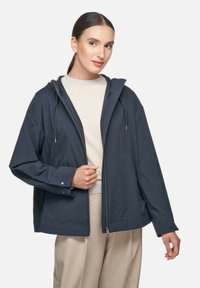 Middenblauwe lichtgewicht jas met een capuchon, voorrits en verstelbare trekkoord. Beschikt over zakken en een ontspannen pasvorm.