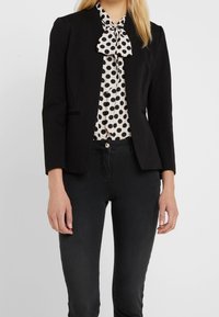 Blazer noir à coupe ajustée, col cranté et deux poches avant, porté sur un chemisier blanc à pois noirs avec un col noué.