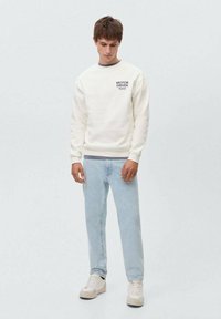 Felpa color crema con scollatura rotonda e testo stampato sul petto, abbinata a jeans azzurri chiari e sneakers bianche.