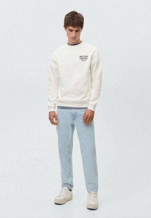 Krämfärgad sweatshirt med rund halsringning och tryckt text på bröstet, kombinerad med ljusblå jeans och vita sneakers.