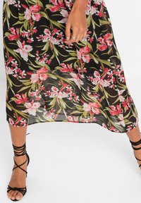 Černé květinové šaty s růžovými a bílými květy, vyrobené z průsvitného materiálu. Mají vzdušný střih a jsou kombinovány s černými sandály se šňůrkami.