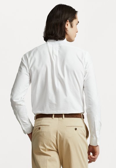 Sötét hajú férfi fehér hosszú ujjú inget visel, amely be van tűrve bézs nadrágba barna övvel, hátulról nézve.