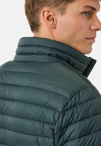 Giacca imbottita di color teal scuro con colletto alto, caratterizzata da un design a cuciture orizzontali e logo ricamato "SWEDEMOUNT" sulla spalla.