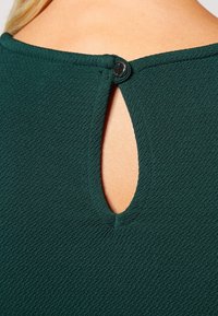 Zelená texturovaná látka s výřezem na zádech, zajištěným knoflíkem na výstřihu. Jednoduchý, čistý design.