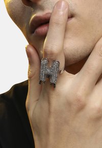 Sølv signetring formet som et "M," prydet med klare rhinsten og grønne accentsten, båret på en finger mod en neutral baggrund.