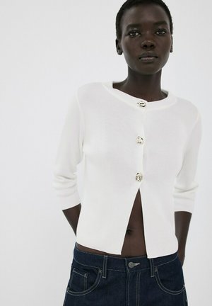 Persona de piel oscura que lleva un suéter blanco corto con tres botones decorativos y jeans azul oscuro sobre un fondo blanco liso.