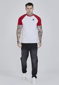 Lühikeste varrukatega raglan-särk punaste varrukatega ja valge kehaga, millel on väike must logo. Kombineeritud mustade teksade ja valgete spordijaladega.