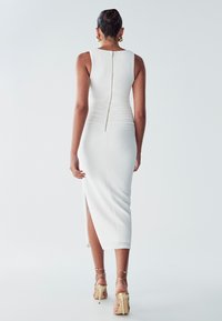 Abito bianco aderente con texture a coste, zip sulla schiena e spacco laterale. Completato da decollete beige con fasce.