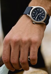 Reloj metálico plateado con una correa de tela azul marino. Presenta una esfera negra con números arábigos blancos y una pantalla de fecha. Correa texturizada.