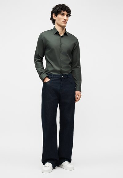 Νεαρός άνδρας με σγουρά μαλλιά, φοράει ένα σκούρο πράσινο πουκάμισο με κουμπιά, φαρδιά σκούρα μπλε τζιν, λευκά αθλητικά παπούτσια, στέκεται με το ένα χέρι στην τσέπη.