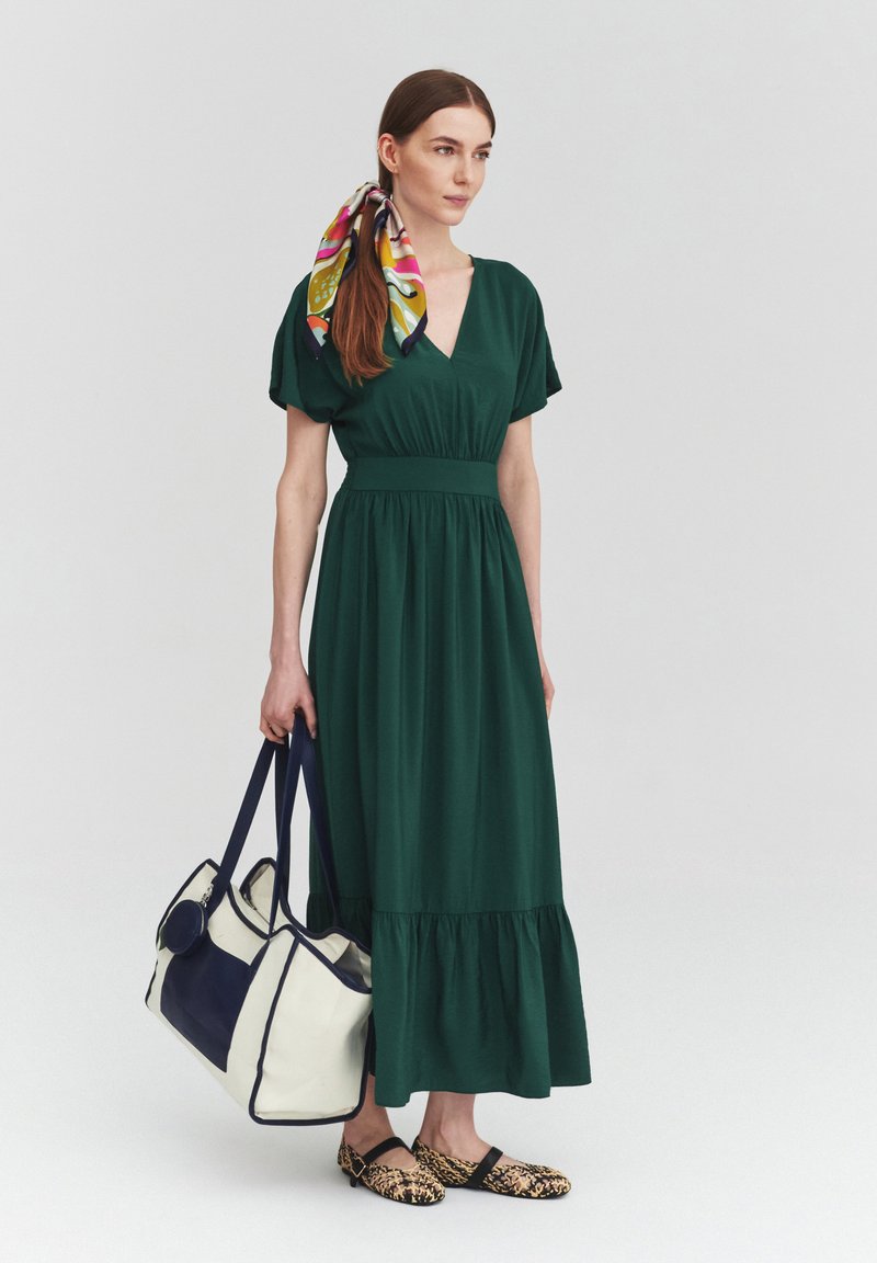 Femme portant une robe midi verte, un foulard coloré dans les cheveux, des ballerines imprimé léopard et tenant un grand sac cabas blanc et bleu marine.