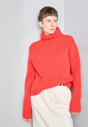 Flauschiger Strickpullover in lebhaftem Koralle mit hohem Rollkragen, gerippter Textur und überschnittenen Schultern, kombiniert mit einem hellen Rock.