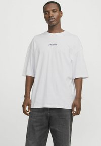 Camiseta blanca de gran tamaño hecha de algodón, con mangas cortas y un logo púrpura de "ARCADIA" impreso en el frente.