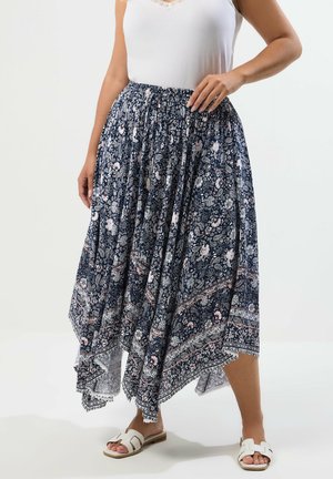 Kobieta w białym, bezrękawnikowym topie, granatowej asymetrycznej spódnicy w kwiaty oraz białych sandałach z odkrytymi palcami stoi na jednolitym tle.