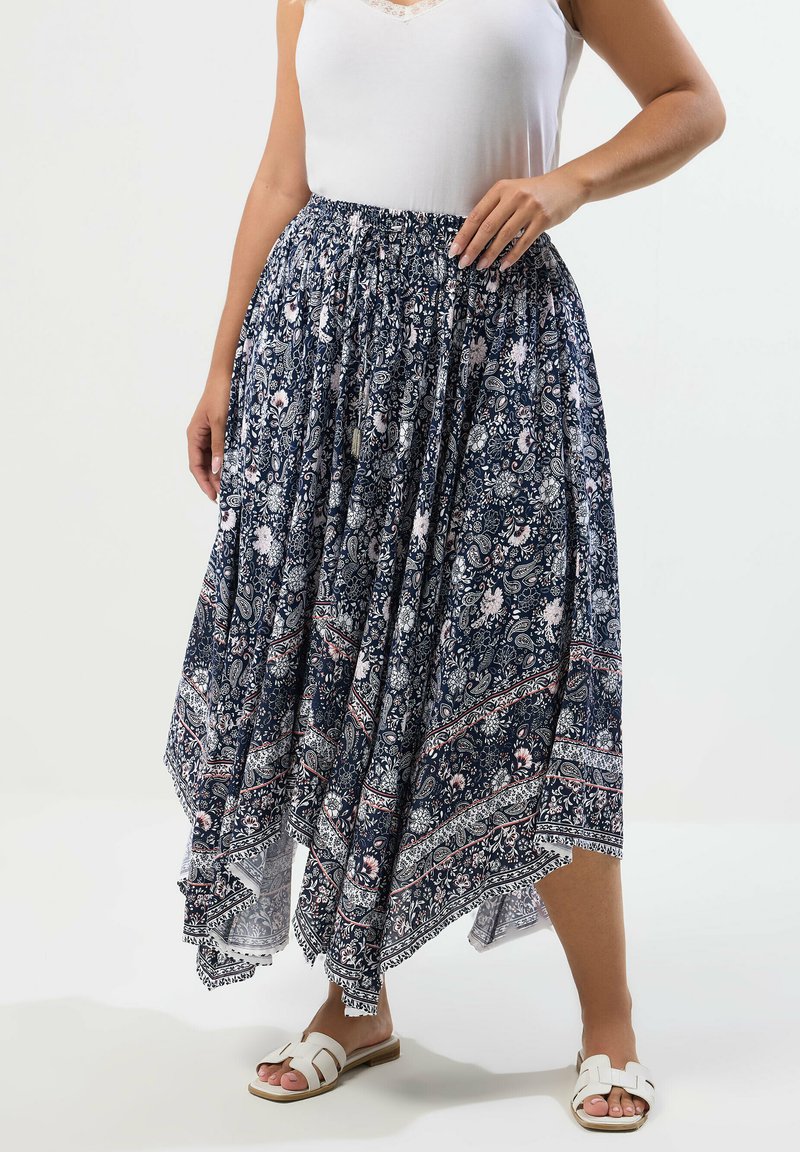 Kvinde iført hvid ærmeløs top, marineblå asymmetrisk nederdel med blomsterprint og hvide sandaler med åben tå, stående mod en ensfarvet baggrund.