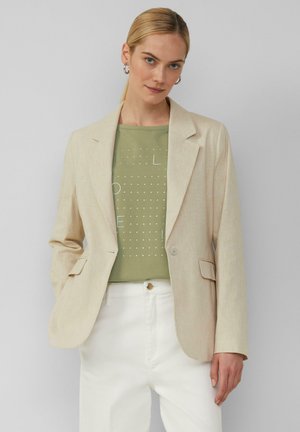 Blazer en lin beige avec revers notés, fermeture à un bouton et deux poches avant, porté sur un top à motifs verts et un pantalon blanc.