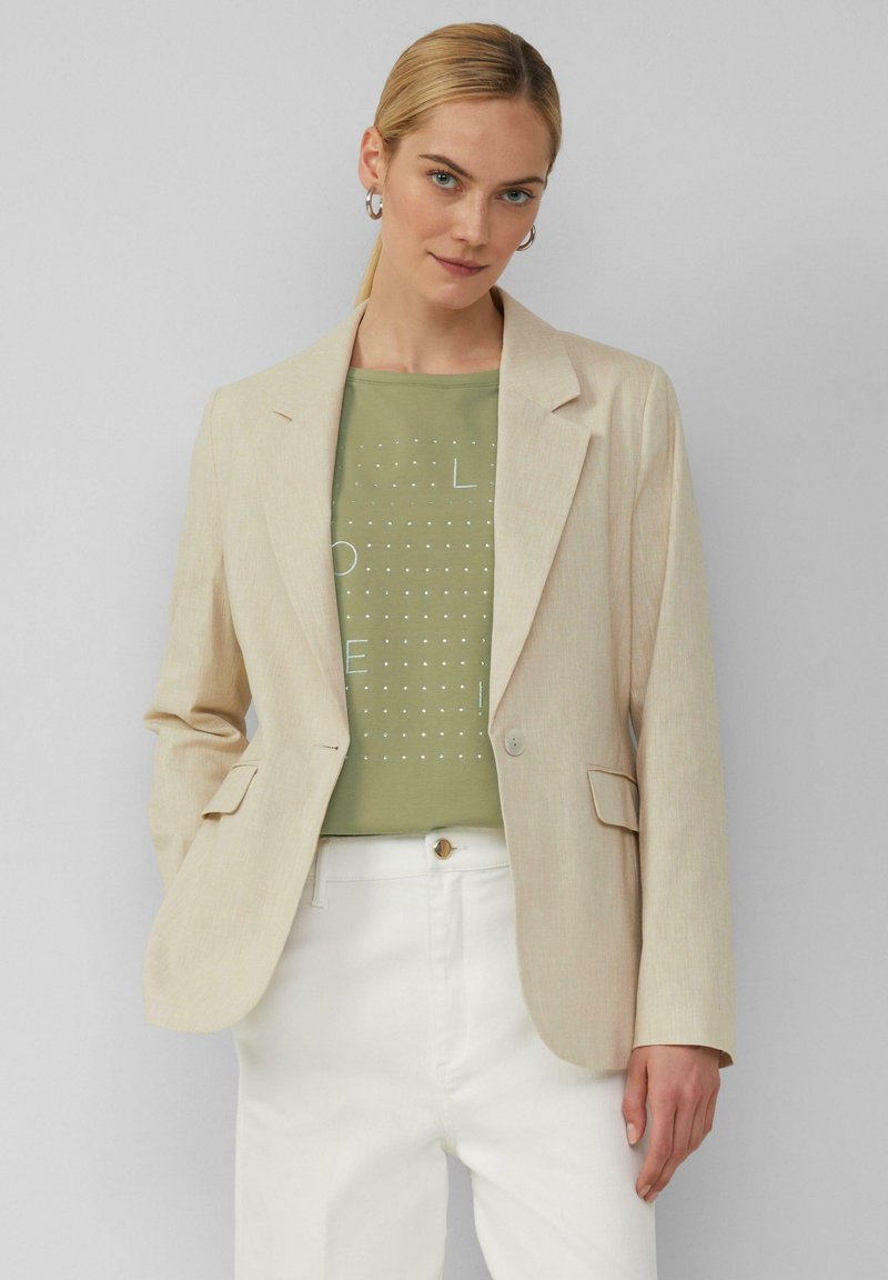 Beige Leinenblazer mit Reverskragen, einreihiger Knopfverschluss und zwei Vorderaschen, getragen über einem grün gemusterten Oberteil und weißen Hosen.