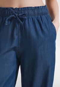 Osoba ubrana w ciemnoniebieskie dżinsy z elastycznym pasem i zawiązanym sznurkiem, ręka w prawej kieszeni, tors częściowo widoczny.