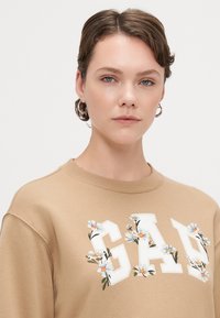 Sweatshirt castanho com o texto "GAP" em branco, adornado com desenhos florais. Textura suave e decote redondo; ajuste casual.