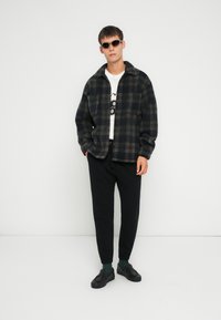 Jachetă din flanel cu carouri în nuanțe de albastru închis și gri, asortată cu un tricou alb cu imprimeu grafic negru, pantaloni jogger negri și adidași negri.