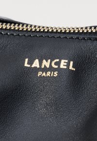 Črn usnjen material z teksturirano površino, z zlatim odtisnjenim logotipom z napisom "LANCEL PARIS" nad zlatim zadrgo.