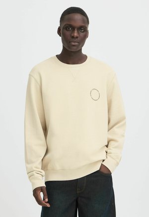Man wearing a beige sweatshirt with circular text on the left chest and dark jeans, standing with one hand in pocket against a white background.
