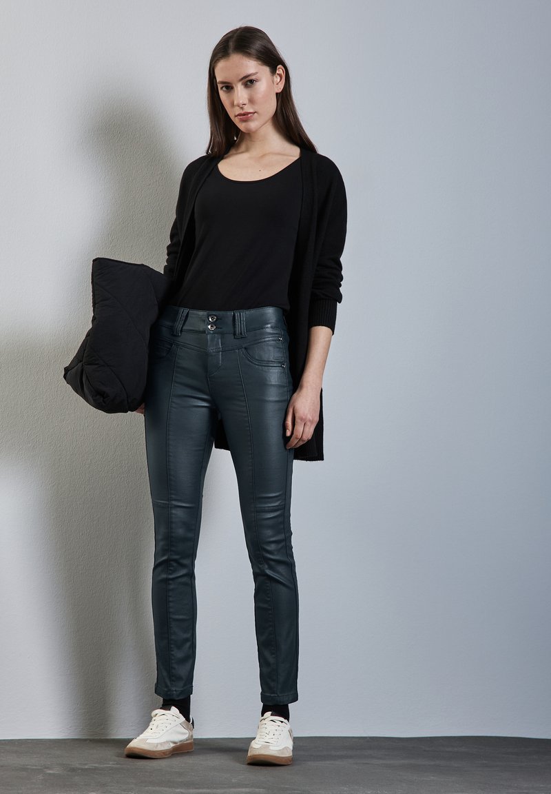 Pantalones ajustados de imitación piel en un tono verde azulado oscuro con dos bolsillos frontales, dos botones en la cintura y una textura suave. Combinados con una blusa negra de manga larga y zapatillas blancas.