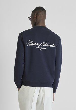 Homme portant un sweat-shirt bleu marine avec le texte blanc « Antony Morato » et le logo au dos, associé à un pantalon blanc, vu de dos.